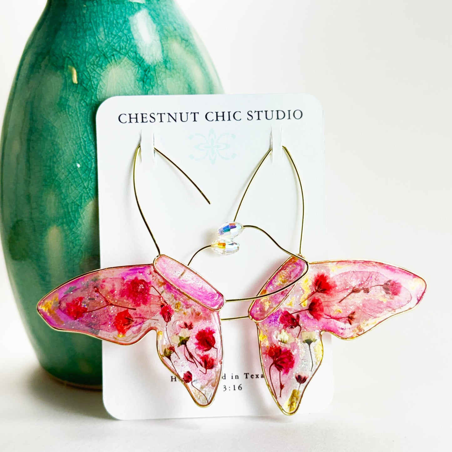 Butterfly-shaped earrings with pink pressed flower wings featuring floral designs on a white card with 'Chestnut Chic Studio' branding, against a white background.