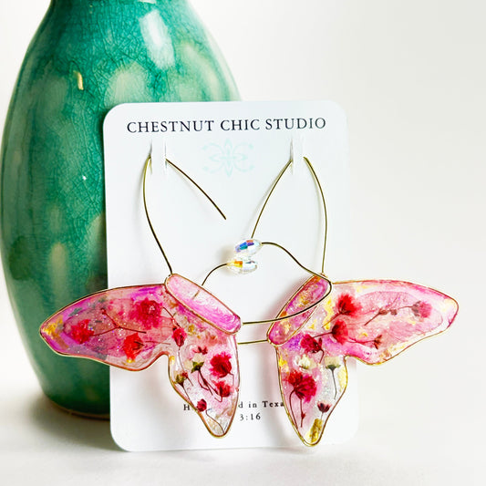 Butterfly-shaped earrings with pink pressed flower wings featuring floral designs on a white card with 'Chestnut Chic Studio' branding, against a white background.